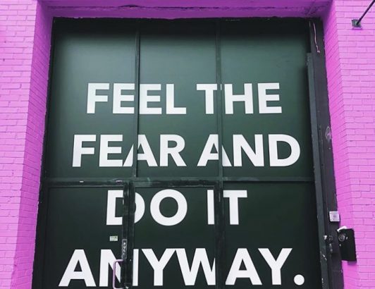 Motivational wall sign reading feel the fear and do it anyway on green doors framed by bright pink brick wall.