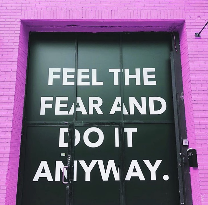Motivational wall sign reading feel the fear and do it anyway on green doors framed by bright pink brick wall.