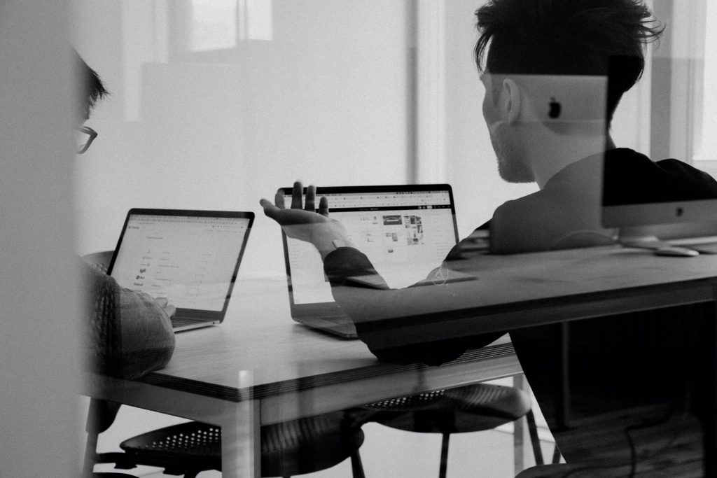 Black and white photo of two colleagues discussing work at table with laptops in modern office, viewed through glass.