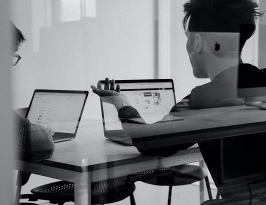 Black and white photo of two colleagues discussing work at table with laptops in modern office, viewed through glass.