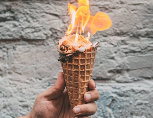 Hand holding ice cream cone with scoop on fire against stone wall background, creating surreal contrast between heat and dessert.