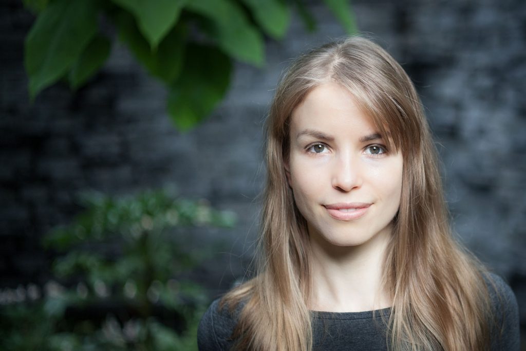 Natural light portrait of woman with long hair standing outdoors against stone wall and greenery, conveying calm and approachability.