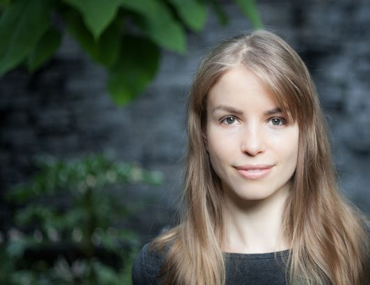 Natural light portrait of woman with long hair standing outdoors against stone wall and greenery, conveying calm and approachability.