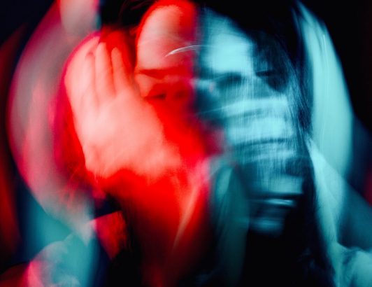 Abstract portrait with heavy motion blur showing a person’s face and hands streaked in red and blue light against a dark background, creating a sense of movement and intensity.