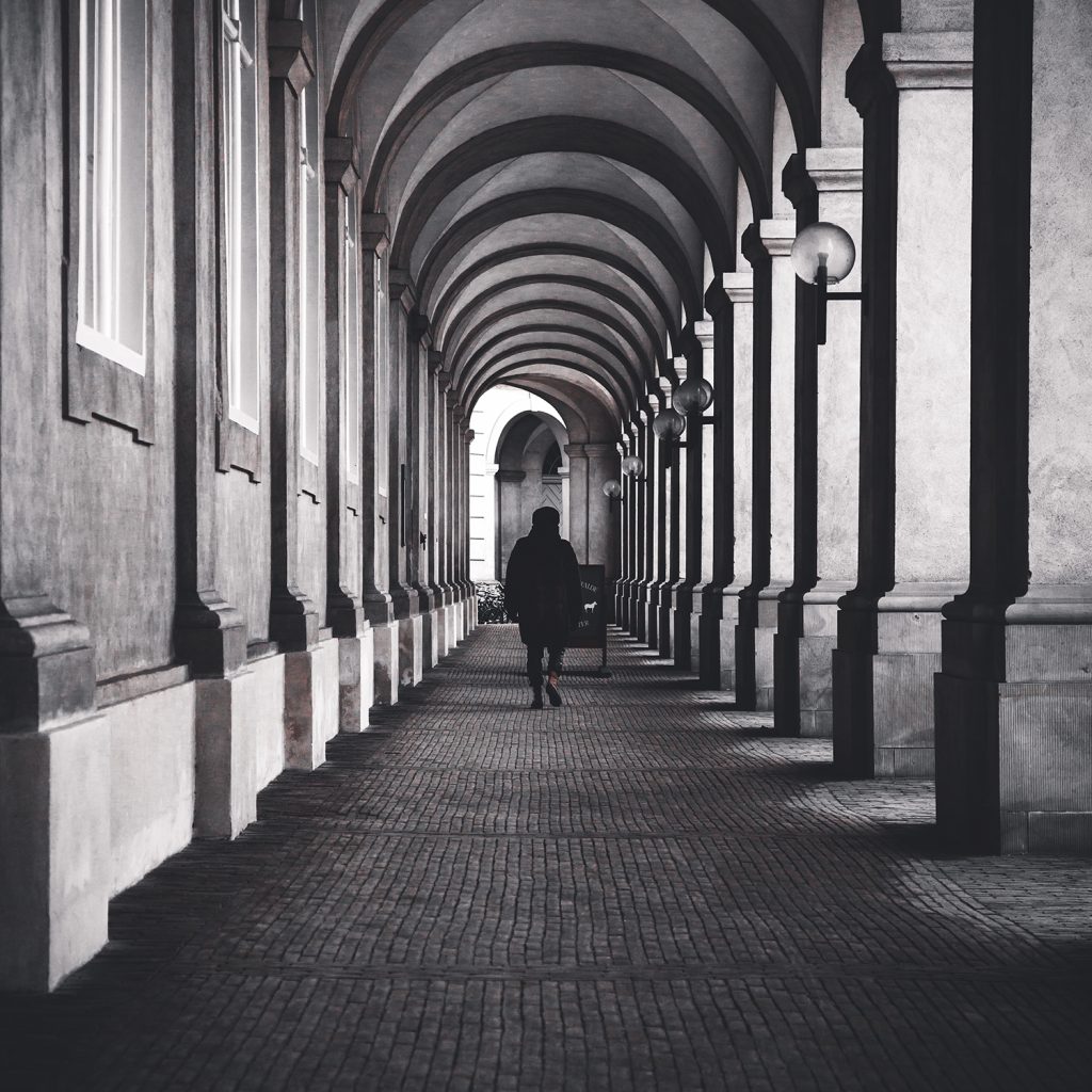 Black and white arched corridor with repeating columns and a lone figure walking, emphasizing symmetry, depth, and quiet solitude.