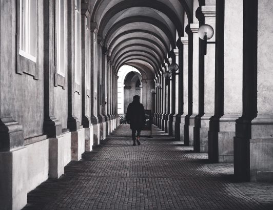 Black and white arched corridor with repeating columns and a lone figure walking, emphasizing symmetry, depth, and quiet solitude.