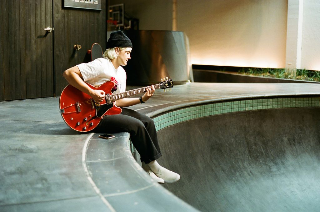 Person sitting on skate park edge playing red electric guitar indoors, blending music, creativity, and urban lifestyle.