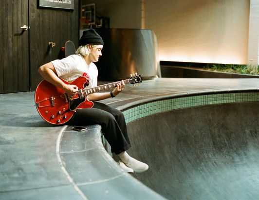 Person sitting on skate park edge playing red electric guitar indoors, blending music, creativity, and urban lifestyle.