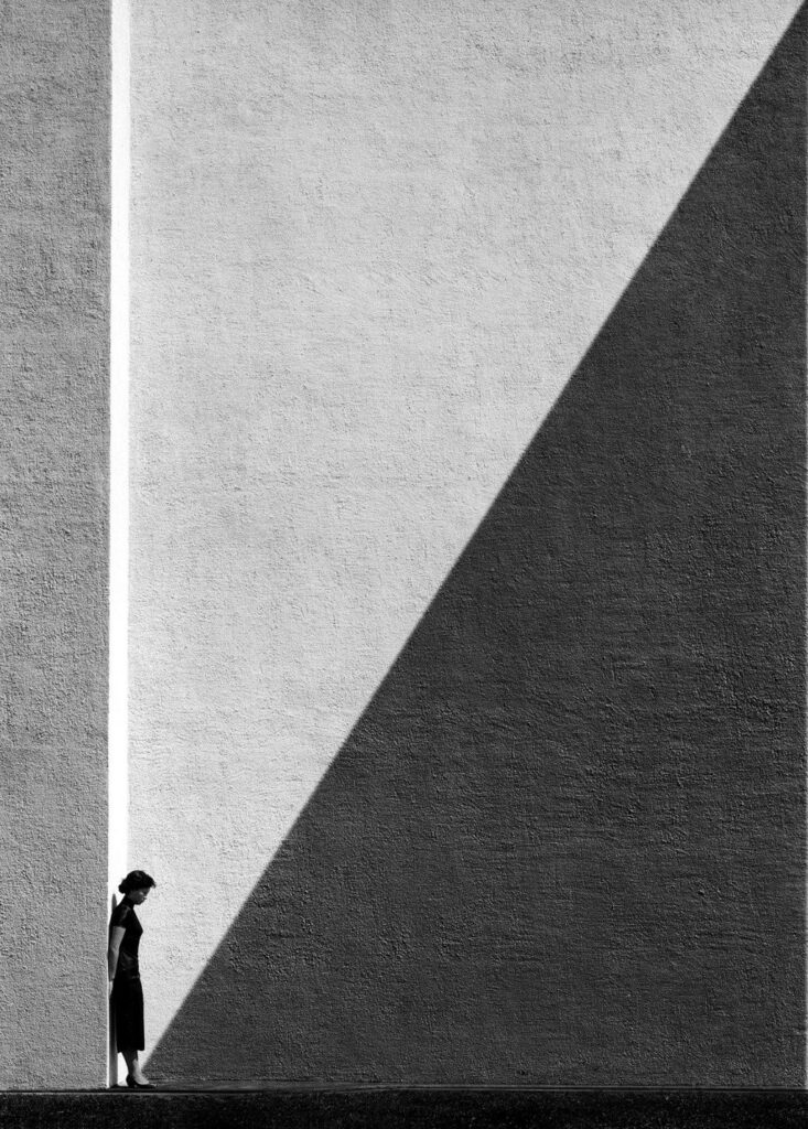 Black and white minimalist scene of woman standing against textured wall with dramatic light and shadow, conveying solitude and reflection.