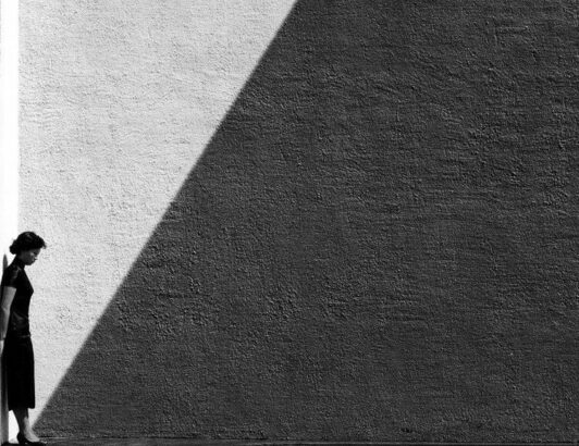 Black and white minimalist scene of woman standing against textured wall with dramatic light and shadow, conveying solitude and reflection.