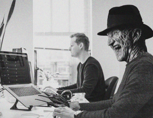 Horror-style figure wearing a hat working at a laptop beside an office coworker in a modern workspace.