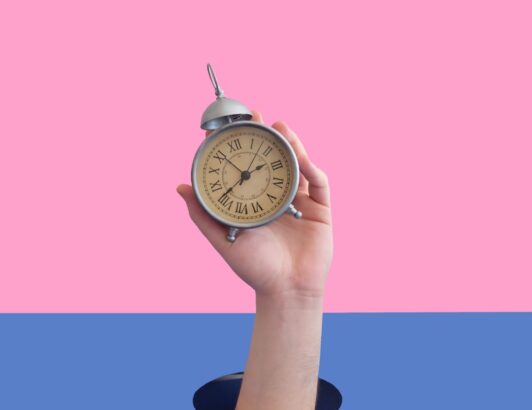 Hand emerging from surface holding vintage alarm clock against pink and blue background, symbolizing time management, urgency, and deadlines.