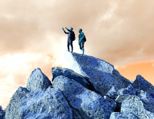 Two hikers standing atop jagged blue-tinted rocks under a dramatic orange sky, surveying the landscape.