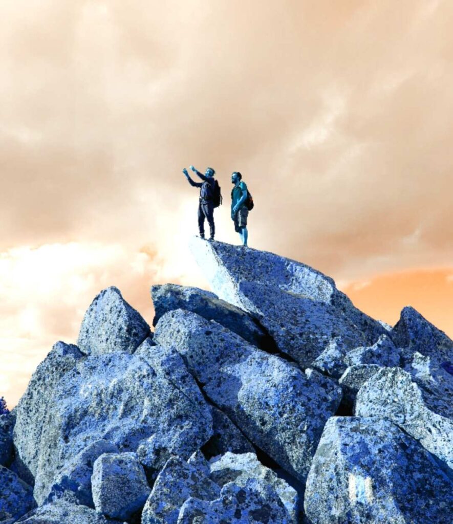 Two hikers standing atop jagged blue-tinted rocks under a dramatic orange sky, surveying the landscape.