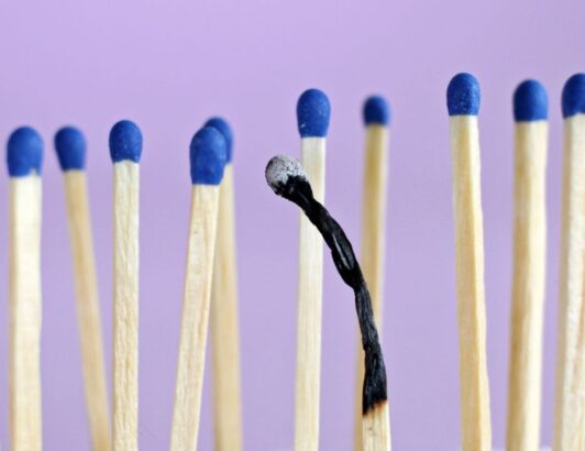Single burnt matchstick surrounded by unlit blue-tipped matches on a purple background, symbolizing burnout.