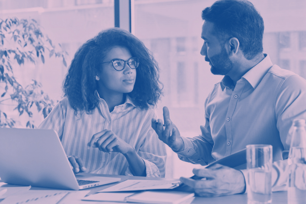 Two colleagues in discussion at a desk with a laptop and documents, collaborating on a project in a bright office.