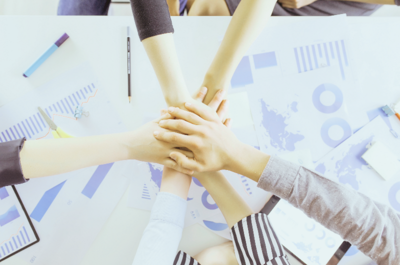Team members stacking hands over desk with charts and graphs, symbolizing teamwork, collaboration, and shared business goals.