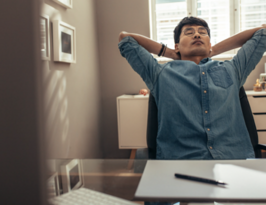 Man leaning back with hands behind head at desk in home office, conveying relaxation, stress relief, or work break.