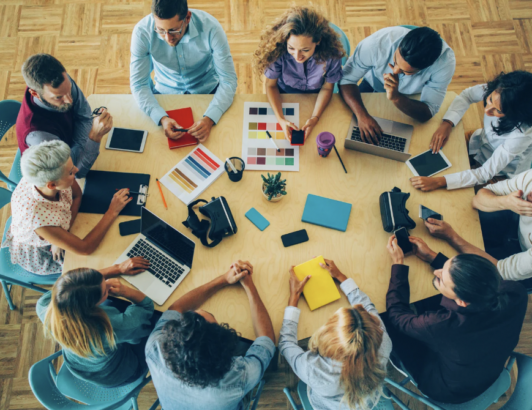 Overhead view of diverse team collaborating around table with laptops, tablets, and design materials during group meeting.