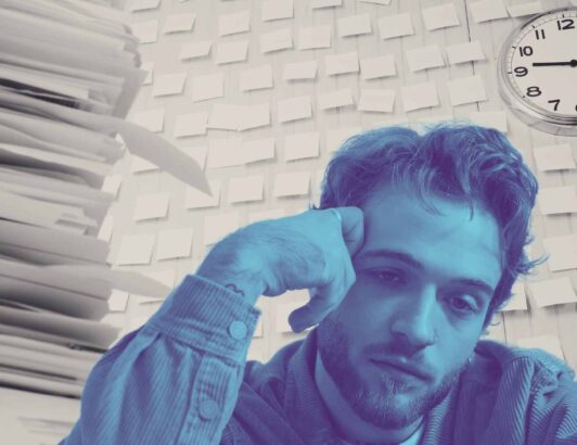 Man sitting at desk with stacks of paperwork, sticky notes on wall, and clock overhead, representing overwhelm, deadlines, and work stress.