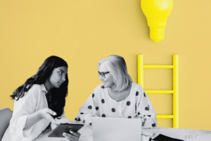 Two women sitting at a desk working, with a ladder and a target in the background.